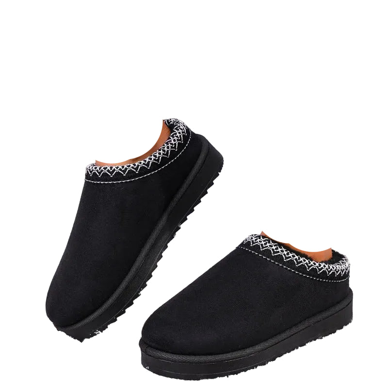 Black slip-on winter slippers with white zigzag trim, shown on a person's feet outdoors in snow.