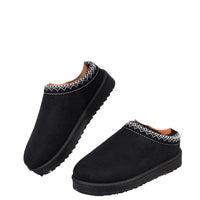 Black slip-on winter slippers with white zigzag trim, shown on a person's feet outdoors in snow.
