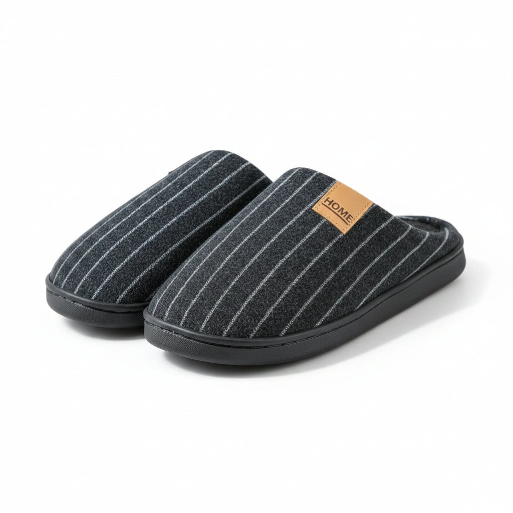 A person holds up a pair of dark gray pinstriped indoor slippers with a soft black lining and a tan leather-like patch that says HOME. The background is a brown curtain.