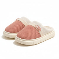 A pair of slip-on indoor slippers with a pink ribbed knit upper, cream elastic band, and cream plush lining, displayed on a wooden stand over a light rug.