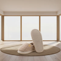 A pair of beige sherpa slip-on slippers shown on a light cream background, with one slipper displaying its textured sole and the other showing the plush interior and a small tan brand tag.