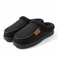 A pair of black slip-on mule slippers with a plush dark gray sherpa lining and a small orange brand tag reading 'Pimochila', displayed on a wooden surface next to a blue box.