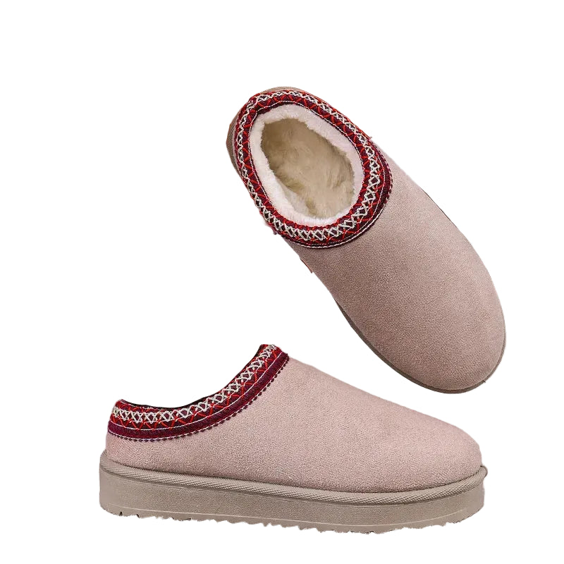 A person wearing a brown fuzzy sock with embroidered eyes is holding up a beige slip-on winter boot with a red and white patterned trim, set against a snowy outdoor backdrop.