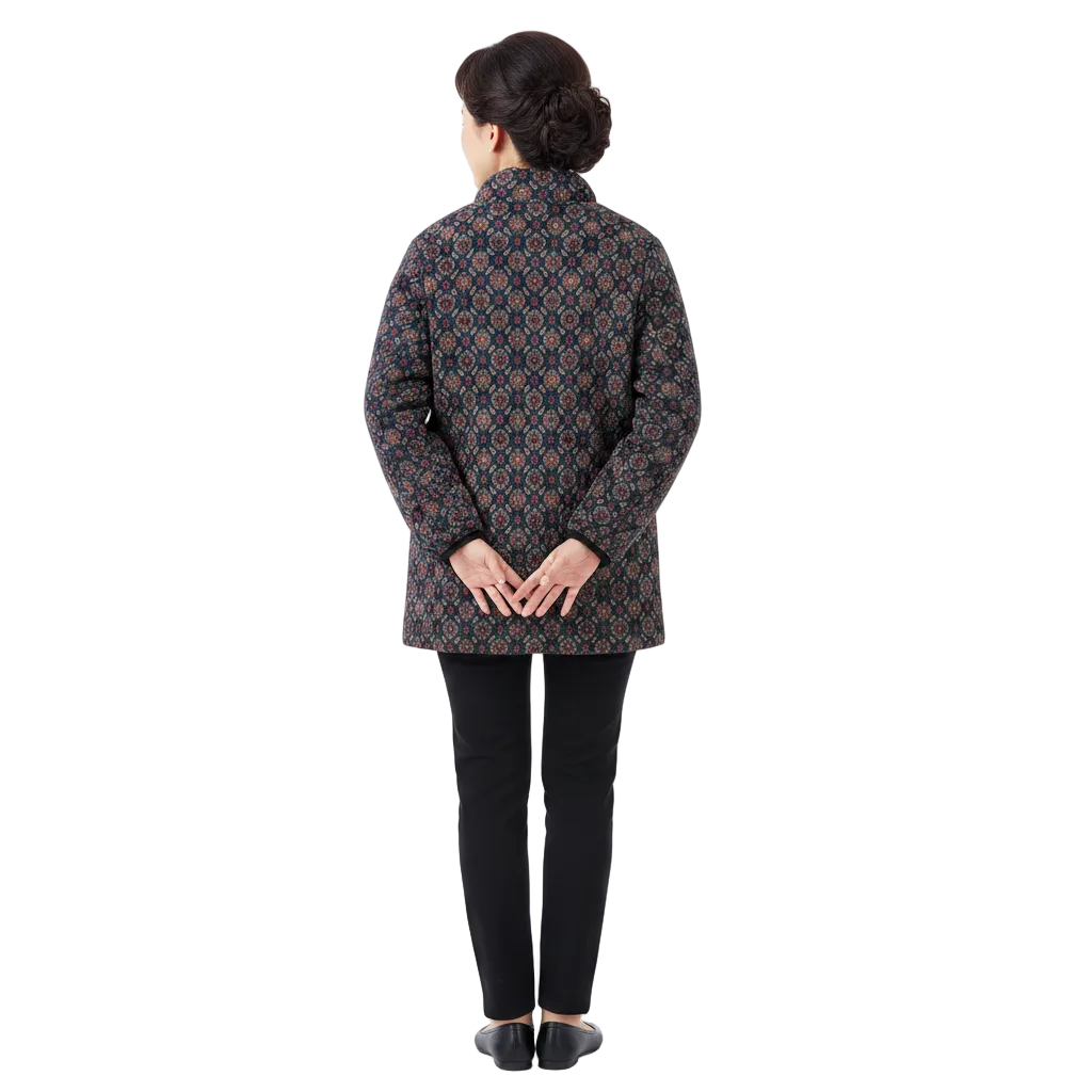 A woman viewed from the back wearing a dark quilted jacket with an intricate geometric pattern in blue, orange, and red, paired with black pants and black shoes, against a plain white studio background.