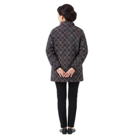 A woman viewed from the back wearing a dark quilted jacket with an intricate geometric pattern in blue, orange, and red, paired with black pants and black shoes, against a plain white studio background.