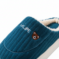 A close-up of a teal blue ribbed fleece slipper with a white sherpa lining, featuring an embroidered white brand name and a small brown bear face on the side.
