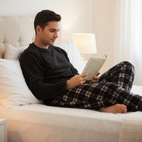 A man relaxes on a white bed reading a book, wearing a black long-sleeve shirt and red, black, and white plaid pajama pants in a softly lit bedroom.
