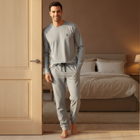 A man wearing a light gray long-sleeve pajama set with matching jogger pants stands barefoot in a doorway, with a neatly made bed and warm bedroom lighting in the background.