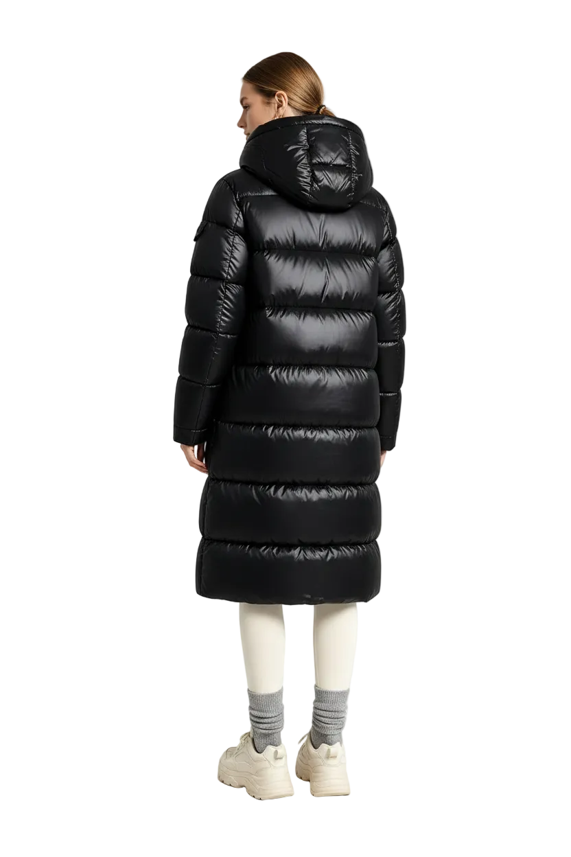 A model viewed from the back wearing a long, black, puffer-style hooded parka coat, paired with cream leggings, gray socks, and beige sneakers, set against a minimalist white studio background.