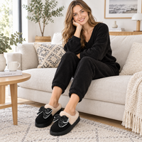 A woman sits on a light beige sofa in a cozy living room, wearing a matching black lounge set with fluffy cream lining and black slip-on mule slippers with bow details. The scene includes a side table with a mug and book, plants, and home decor.