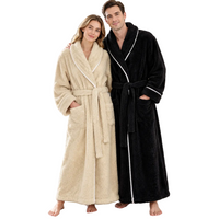 A man and a woman standing barefoot in a bright bedroom, each wearing a plush bathrobe. The woman's robe is cream/beige with white piping, and the man's robe is black with white piping.