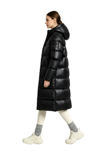 A woman modeling a long black puffer coat in a studio setting. She is shown in profile walking, wearing cream leggings, gray socks, and chunky off-white sneakers.
