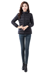 A woman modeling a black puffer jacket with a high collar and front zipper, paired with dark blue skinny jeans and black ankle boots, against a plain white studio background.