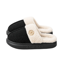 A pair of slip-on mule slippers with a black textured upper, cream sherpa lining, and a black sole embossed with the brand name COMFORT. A small circular beige logo patch is visible on the front strap.