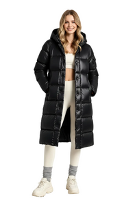 A woman modeling a long black puffer coat in a bright, minimalist studio setting. The coat is glossy and features horizontal quilting, a hood, and a front zipper closure.
