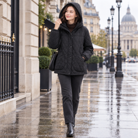A woman walking on a wet city street wearing a black quilted hooded jacket, dark pants, and black ankle boots, with a blurred classical building in the background.