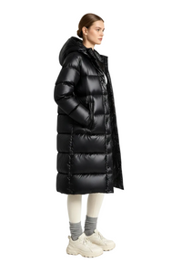 A model wearing a long black puffer coat with a high collar, paired with cream-colored pants, gray socks, and beige sneakers, standing in a bright white studio.