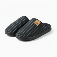 A pair of black slip-on slippers with thin white vertical pinstripes, featuring a tan leather brand patch on the side that reads 'HOME'.