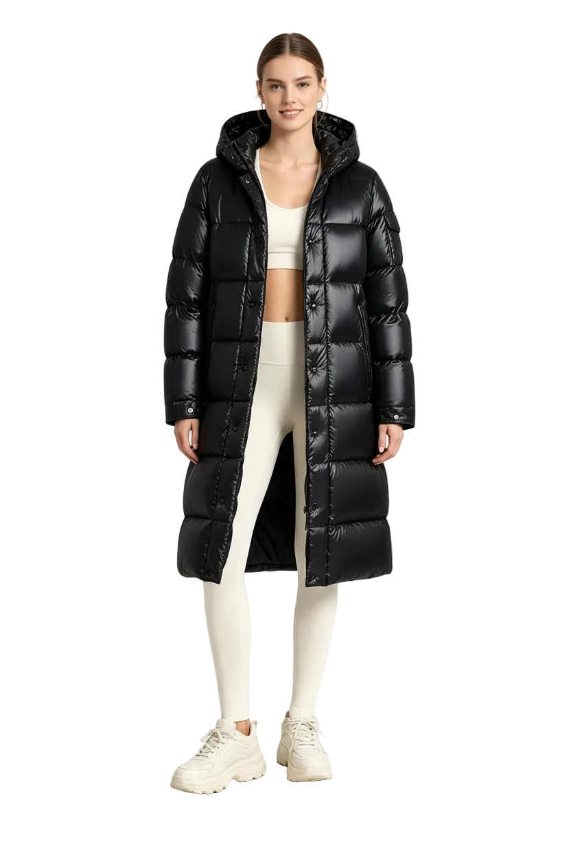 A woman stands in a studio wearing a long black puffer coat with a hood, paired with cream leggings, a matching crop top, and cream sneakers.