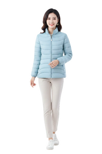 A woman modeling a light blue puffer jacket with a stand-up collar, paired with beige pants and white sneakers, against a plain white studio background.