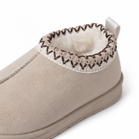 A close-up of a beige slip-on slipper with a white faux-fur lining, a white elastic trim, and dark brown zigzag stitching along the collar.