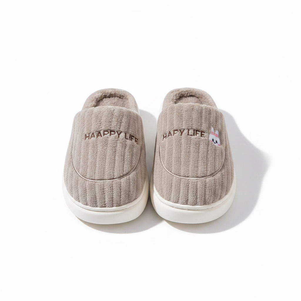 A pair of light taupe ribbed slip-on slippers with white soles, featuring 'HAPPY LIFE' embroidered on the side and a small cartoon bunny face on the right slipper.