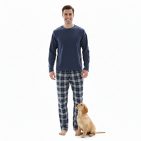 A man wearing a navy blue long-sleeve shirt and blue-and-white plaid pajama pants stands barefoot on a white background, with a golden retriever puppy sitting at his feet.