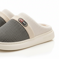 A close-up studio shot of a pair of slip-on mule slippers. The slippers feature a textured dark gray upper, a ribbed cream elastic band with a black, red, and white 'YOTMAG' brand patch, and a thick cream-colored sole.