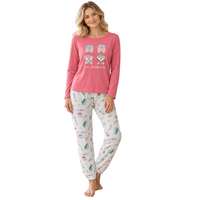 A woman stands barefoot in a bright living room wearing a pink long-sleeve pajama top with a small graphic print and white patterned pajama pants. The room includes a light beige armchair with a green throw blanket, a wooden side table with a plant, and a bookshelf in the background.