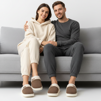 A man and a woman sitting on a light gray couch, wearing matching loungewear sets and brown slip-on slippers with white trim.