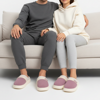 A couple sitting on a light beige couch wearing matching cozy loungewear sets and pink-and-cream slip-on slippers. The man wears a dark gray long-sleeve top and jogger pants, and the woman wears a cream hoodie and light gray leggings.