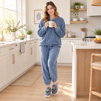 A woman stands in a bright, modern kitchen wearing a matching blue loungewear set with a sweatshirt and jogger pants, holding a white mug. The set features a heathered blue texture, and she is wearing blue slip-on slippers with cartoon bear patches.