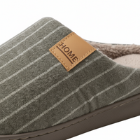 A close-up of a slip-on mule slipper in textured gray fabric with thin white stripes, featuring a tan faux-leather brand patch that reads HOME and a soft fleece lining.