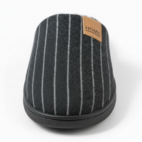 A single slip-on house slipper shown from a rear three-quarter view on a white background. The slipper features a dark gray textured fabric upper with vertical white pinstripes, a tan leather brand patch reading 'HOME' on the side, and a black rubber sole.