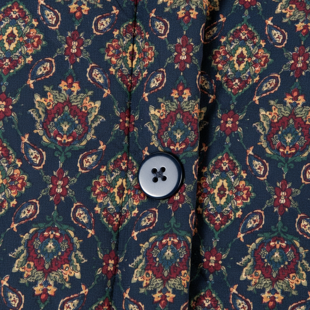 Close-up of a button-front garment with a dark navy base and an ornate floral paisley pattern in red, green, and beige, featuring a single four-button snap closure.
