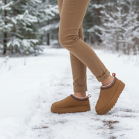 A person wearing tan winter slippers with red trim and brown platform soles, standing in a snowy forest path.