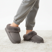 A person wearing gray slip-on slippers with fluffy faux-fur trim, standing on a white textured rug. The person is also wearing gray jogger pants.