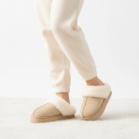 A person wearing beige slip-on slippers with white faux-fur trim, paired with cream jogger pants, standing on a white textured rug against a white background.