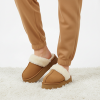 A person wearing tan fleece-lined slip-on slippers with platform soles, paired with tan jogger pants, standing on a white shag rug against a white studio background.
