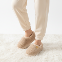A person wearing beige sherpa slip-on slippers with cream jogger pants, standing on a white shag rug.