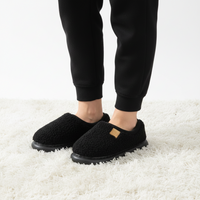 A person wearing black fleece slip-on slippers with black jogger pants, standing on a white shag rug.