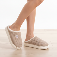 A person wearing beige ribbed slip-on house slippers with white faux-fur trim and white soles, standing on a light wood floor against a white wall.