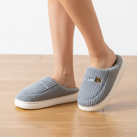 A person wearing light gray ribbed slip-on house slippers with white soles, standing on a light wood floor against a white wall.