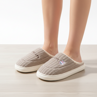 A person wearing taupe ribbed slip-on slippers with white faux-fur lining and a small embroidered cartoon face on the front, standing on a light wood floor against a white wall.