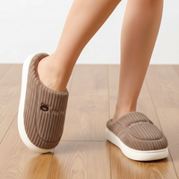 A person wearing brown ribbed slip-on house slippers with white soles, standing on a light wood floor against a white wall.