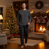 A man wearing a dark gray long-sleeve pajama top and dark gray plaid pajama pants stands barefoot in a cozy living room decorated for Christmas, with a lit fireplace, stockings, and a decorated tree in the background.