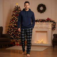 A man wearing a navy blue pullover hoodie and matching navy plaid pajama pants stands barefoot in a cozy, decorated living room with a Christmas tree, fireplace, and wreath.