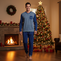 A man wearing a blue and navy striped long-sleeve henley pajama set with matching pants stands barefoot in a cozy living room decorated for Christmas, with a lit fireplace, wreath, and decorated tree in the background.