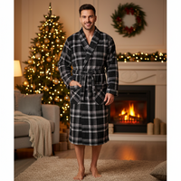 A man wearing a black and white plaid holiday robe stands barefoot in a cozy living room with a lit fireplace, Christmas tree, and wreath in the background.