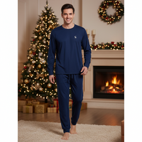 A man wearing a navy blue long-sleeve pajama set with matching pants stands barefoot in a cozy living room decorated for Christmas, with a lit fireplace and a decorated tree in the background.